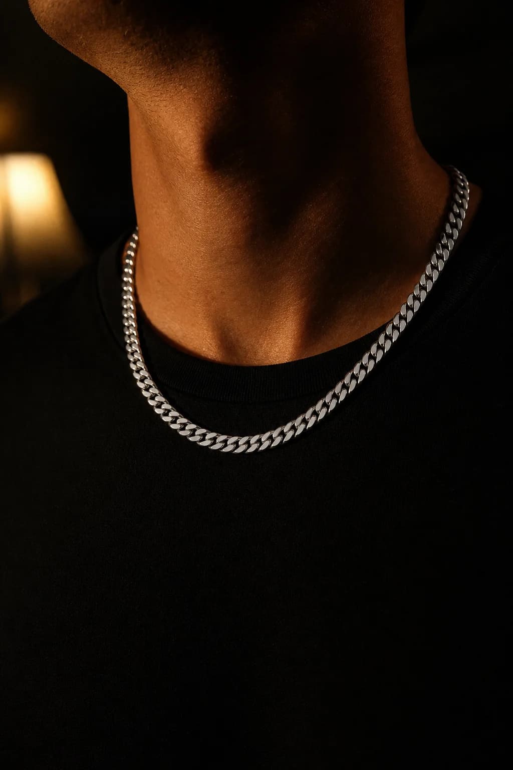 A polished steel Cuban link chain resting on a collarbone over a black t-shirt, lit by one warm lamp